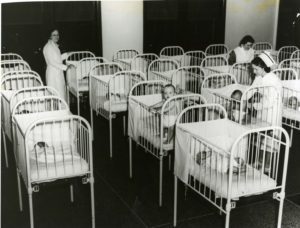 An old fashioned nursery with rows of babies in cribs and three nurses caring for the babies.