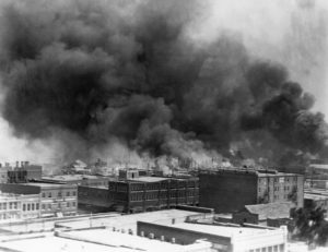 Black and white photo of city buildings, some with thick clouds of smoke billowing from the roofs