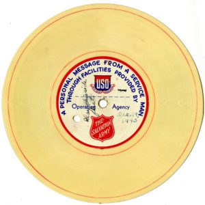 Digital scan of a small yellowish record. The label in the center of the record has blue text which reads: "A personal message from a service man through facilities provided by" Below text is a USO (United Services Organization) logo, text reading "Operating Agency" and a Salvation Army Red Shield logo. Written in ink on the label is text which reads: "Play this side first. Dec. 19, 1943"