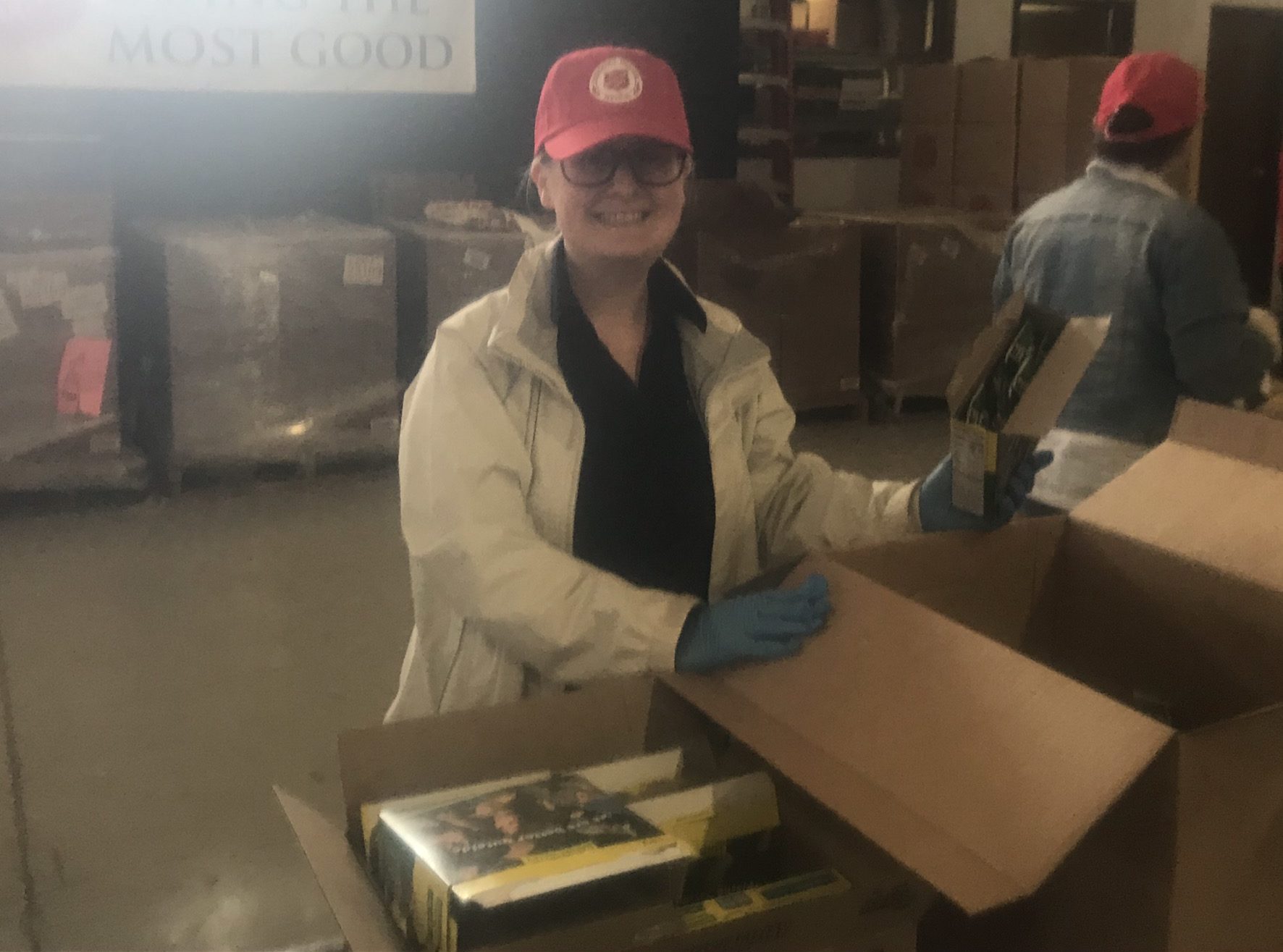 A woman wearing a hat and coat smiles for the camera while packing a cardboard box with food.
