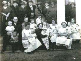 A group of adults, nurses and babies are seated and standing on the front porch of a building.