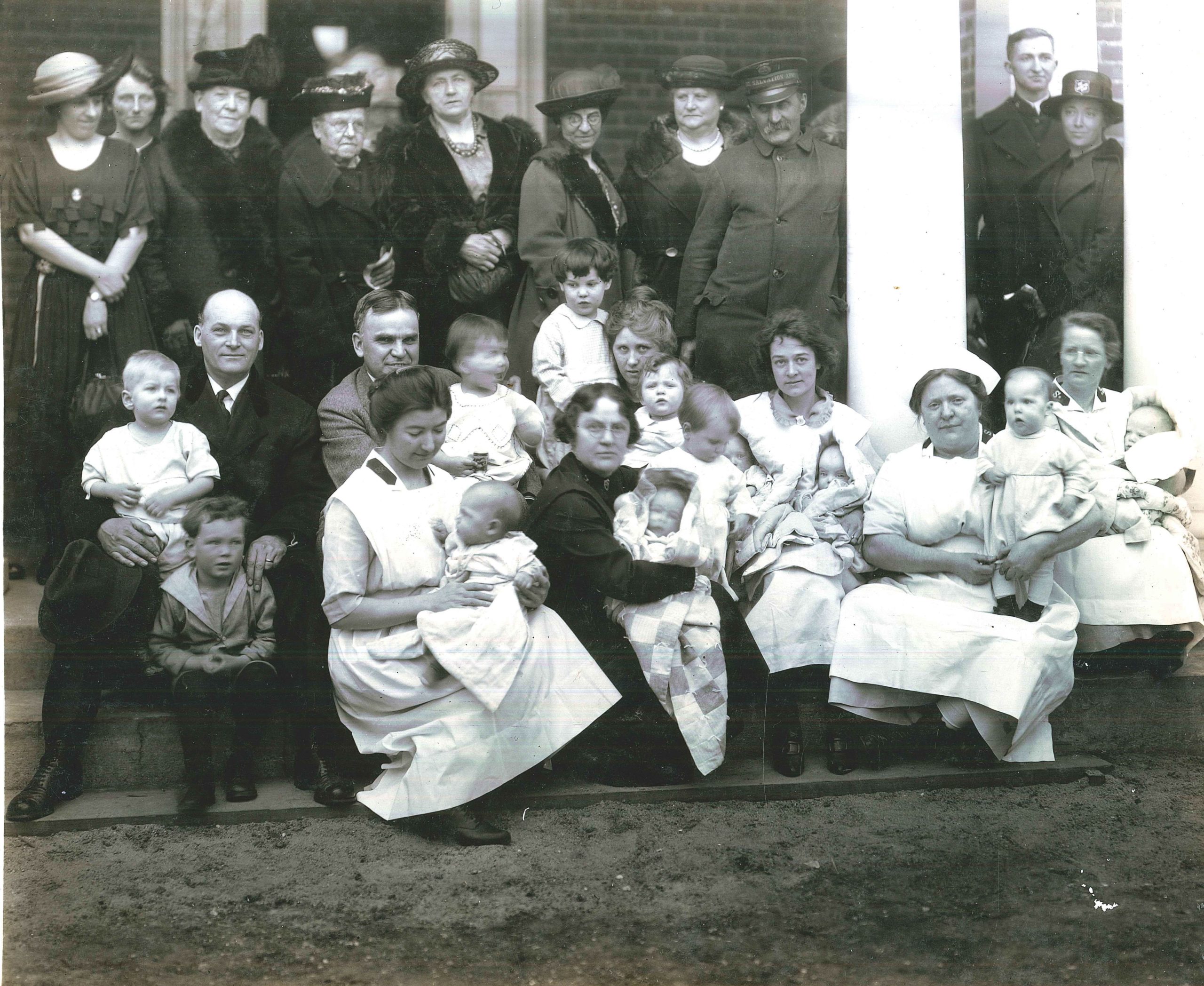 A group of adults, nurses and babies are seated and standing on the front porch of a building.