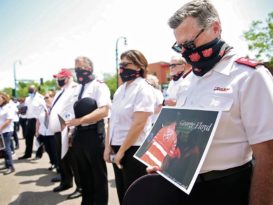 Salvation Army offiers wearing face masks bow their heads in prayer, one holds sign honoring George Floyd