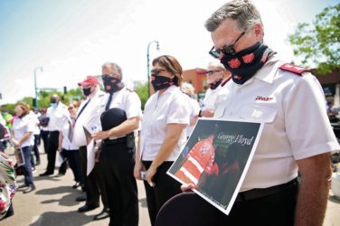 Salvation Army offiers wearing face masks bow their heads in prayer, one holds sign honoring George Floyd
