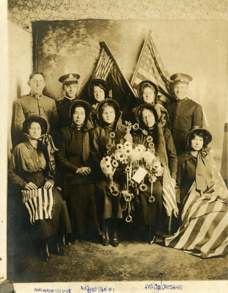 A group of men and women posing with US flags and a wreath decorated with glowers and doughnuts