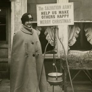 Black and white photo of a woman wearing a cape and soft cloth hat with Red Shield logo patch on front. She stands next to a kettle stand. The placard reads: "The Salvation Army Help us make others happy. Merry Christmas."