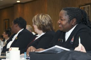 Photograph showing three women seated in a wood paneled conference room. Major Janice Love is on the far right and is smiling.