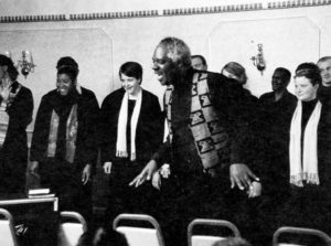 Photo showing a black Salvation Army officer standing in front of a choir. 