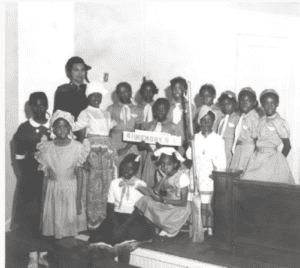 Photograph showing Norma T. Roberts posing with a group of Black girls. The girls are dressed in pioneer style or Sunbeam dresses.