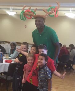 Photograph showing Captain AJ Zimmerman wearing a green polo shirt and an inflatable moose antler ring toss game on his head. A group of children stand with him.