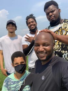 Captain AJ Zimmerman wearing a black shirt poses with five teenage boys. Three of the young men are wearing back facemasks which were used to prevent the spread of the SARS-CoV2 virus in 2020-2021