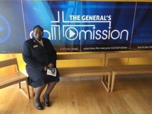 Photograph of Major Katherine Clausell wearing a Salvation Army uniform. She is seated on a wood bench in front of a banner that reads "The General's Call to Mission"