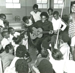 Photograph showing a Lt. Stephen Harper holding a guitar and smiling. He is surrounded by children.