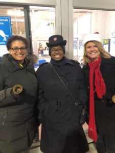 A photograph showing three women wearing winter coats and hats. The woman on the far left, Major Cassandra DeJesus holds a handbell