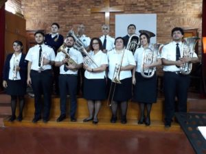 Photo of a Salvation Army brass band. The men and women wear white button down shirts and black pants or skirts. They stand in two rows with their instruments in the alter of the corps.