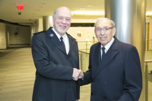 Photograph of two men shaking hands. They wear Salvation Army Soldier uniforms and are standing in an office building