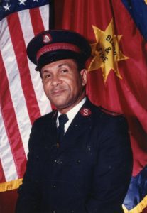 Photograph of Major Allan Wiltshire wearing a Salvation Army uniform. He is standing in front of Salvation Army and United States flags.