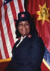 Photograph of Major Marjorie Wiltshire wearing a Salvation Army uniform. She stands in front of Salvation Army and United States flags