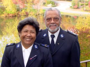 Color photograph of Envoys Roberto and Marina Santos wearing their Salvation Army uniforms. The smiling couple stands in front of a scenic outdoor pond.