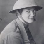 Black and white photograph of a young woman wearing a WWI helmet and military style uniform. She has the strap of a gas mask bag around her neck.