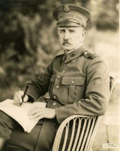 Black and white photograph of an older man with mustache wearing a WWI style military uniform. The man is seated in a chair with pieces of paper on his lap and a pen in his right hand. Trees can be seen in the background.
