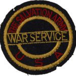 Color photo of a fabric patch. The background fabric is black. Two circles are embroidered with gold tread. Inside the two circles is red text embroidered in red thread that reads "The Salvation Army U S A." Over the circles is more text positioned horizontally, embroidered in gold tread, it reads "War Service."