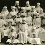 Black and white photo showing a large group of women wearing white fabric caps and smocks over their clothing. The women hold knitting.