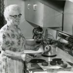 Black and white photo showing a woman with white hair and glasses, wearing a dress and fancy apron. She stands at the stove in her mid-20th century kitchen. She is frying doughnuts