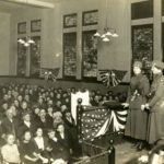 Black and white photo showing people in a chapel or lecture hall. Two Doughnut Girls and two male Salvation Army officers are on the stage.