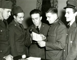 Black and white photograph showing five men, for of them wearing World War II US Army uniforms and one wearing a Salvation Army officer uniform. The man in the center of the image wears a US Army uniform and is holding a microphone. The Salvation Army officer holds a square envelope with text that reads: "A Recordio Message For..."