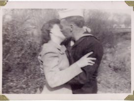 Black and white photo of a United States sailor in uniform kissing a woman in a suit. The couple are outside and landscape plants and shrubs can be seen in the background.