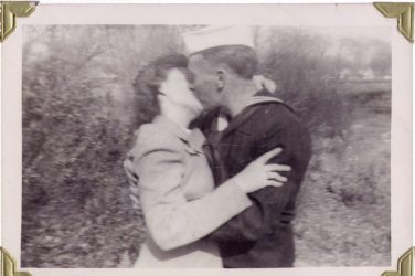 Black and white photo of a United States sailor in uniform kissing a woman in a suit. The couple are outside and landscape plants and shrubs can be seen in the background.