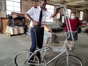 Photograph of an African American Salvation Army officer and woman examining a silver painted tall bike. They are in an abandoned warehouse