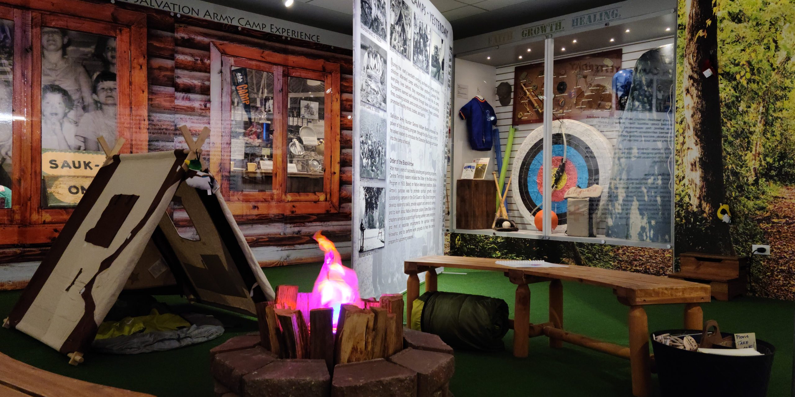 Photograph showing the interior of an exhibit gallery. Exhibit cases can be seen in the background with artifacts inside. One of the cases is decorated to look like a log camp cabin. In the foreground is a tent with artificial fire and bench.