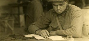 Photograph of a World War I soldier seated at a table with writing material and a dominoes set in front of him. He holds a pen and looks at a blank piece of paper as if deciding what to write in a letter.