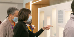 Photograph showing two women wearing facemasks. The women are talking and pointing to a museum exhibit case. Other people can be seen in the backgground.