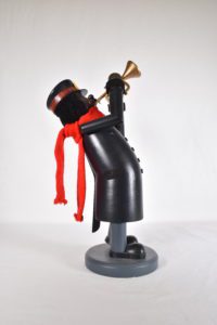 Photograph of a wood nutcracker. It is in the shape of an African American Salvation Army Soldier (church member) playing a cornett. The nutcracker wears a long overcoat, hat and red scarf. 