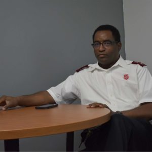 Color photograph of an African American man seated at a table. The man wears a white short-sleeved button down shirt with Salvation Army officer (pastor) insignia.