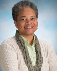 Headshot photograph of a smiling middle aged African American woman wearing a light green button-down shirt, crème color cardigan, and green scarf.