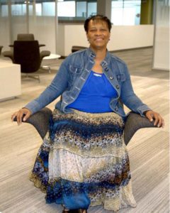 Color photograph of a middle aged African American woman wearing a jean jacket, blue shirt, and long blue skirt. She is seated in a chair with arms.