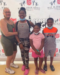 A smiling African American woman and three children standing in front of a banner with Salvation Army and Kroc Center logos printed on it. The woman is middle aged and wears her hair pulled back. She has glasses and wears a cloth facemask below her chin. She is dressed in olive green tank top and shorts. The children, two girls and a boy, also wear casual clothing such as tank tops, t-shirts, shorts, and leggings. The children also wear facemasks below their chin.