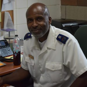 A smiling bald African American man who is seated in a chair, slightly leaning forward in his seat. He wears a white button down shirt with blue Salvation Army envoy epaulets and a gold name tag.