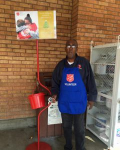 An African American man stands in front of a brick wall and propane cannister container. He wears a blue apron with Salvation Army Red Shield logo and holds a hand bell. He stands next to a Salvation Army red kettle and holder.