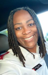 Selfie of a smiling African American woman with long braided hair wearing a white button down shirt with red Salvation Army officer epaulets and a name tag.