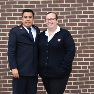 A man and a woman wearing Salvation Army officer uniforms stand in front of a brick wall.