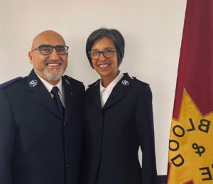 A smiling man and woman wearing Salvation Army uniforms consisting of white button down shirts and navy blue suit coats stand next to a Salvation Army flag on a flagpole. The man is bald with grey goatee and the woman has short dark greying hair. Both wear glasses. 