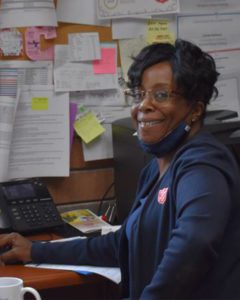 An African American woman seated at a desk. She looks back towards the camera. She is wearing a dark blue facemask which is under her chin, blue shirt, and dark blue hoodie with Salvation Army Red Shield logo