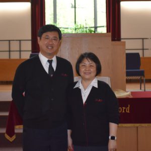 A smiling Korean American couple stand in front of a church altar. Both wear Salvation Army officer uniforms consisting of white button down shirts, navy blue cardigan sweaters, and navy trousers. The man is significantly taller than his wife, being about a head height taller.