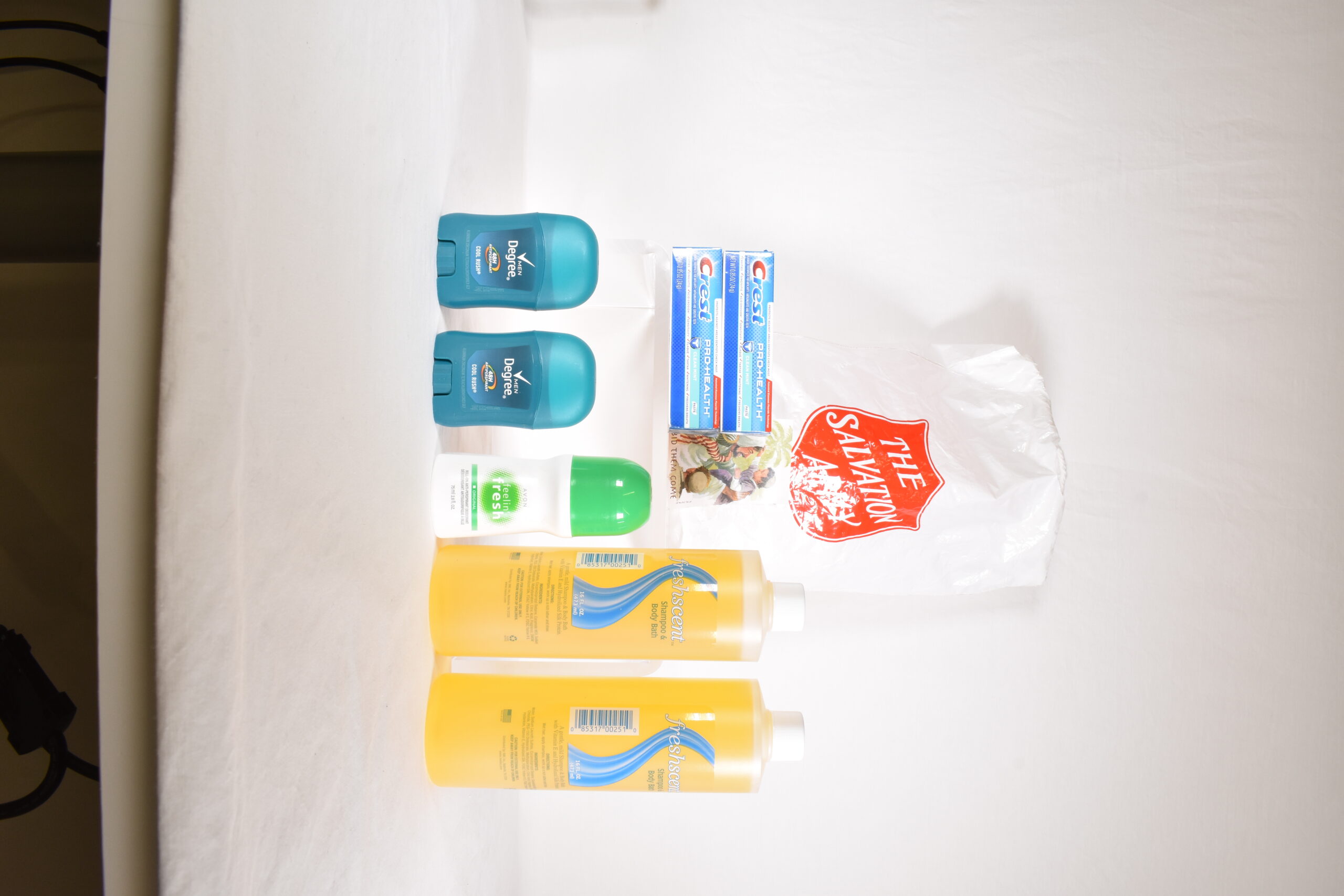 A white plastic drawstring bag with the Red Shield logo on front. Assorted toiletries which are distributed in the bag are arranged around it. These include shampoo, deodorant, and toothpaste.