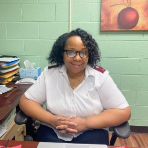 A smiling African American woman with chin length curly hair arranged in a side part, glasses, and white button down shirt with red Salvation Army officer epaulets. She is seated in an office chair and rests her elbows on the chair's arms and interlaces her fingers in front of her. She is seated in an office with aqua painted cinderblock walls.